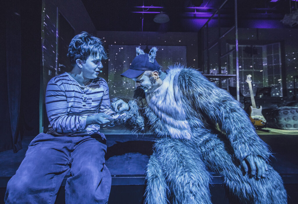 Zdjęcie ze spektaklu Preskot. Zdjęcie zabarwione na niebiesko. Młody aktor siedzi obok aktora w stroju kota i częstuje go czymś. W tle świecą małe światełka jak świetliki.