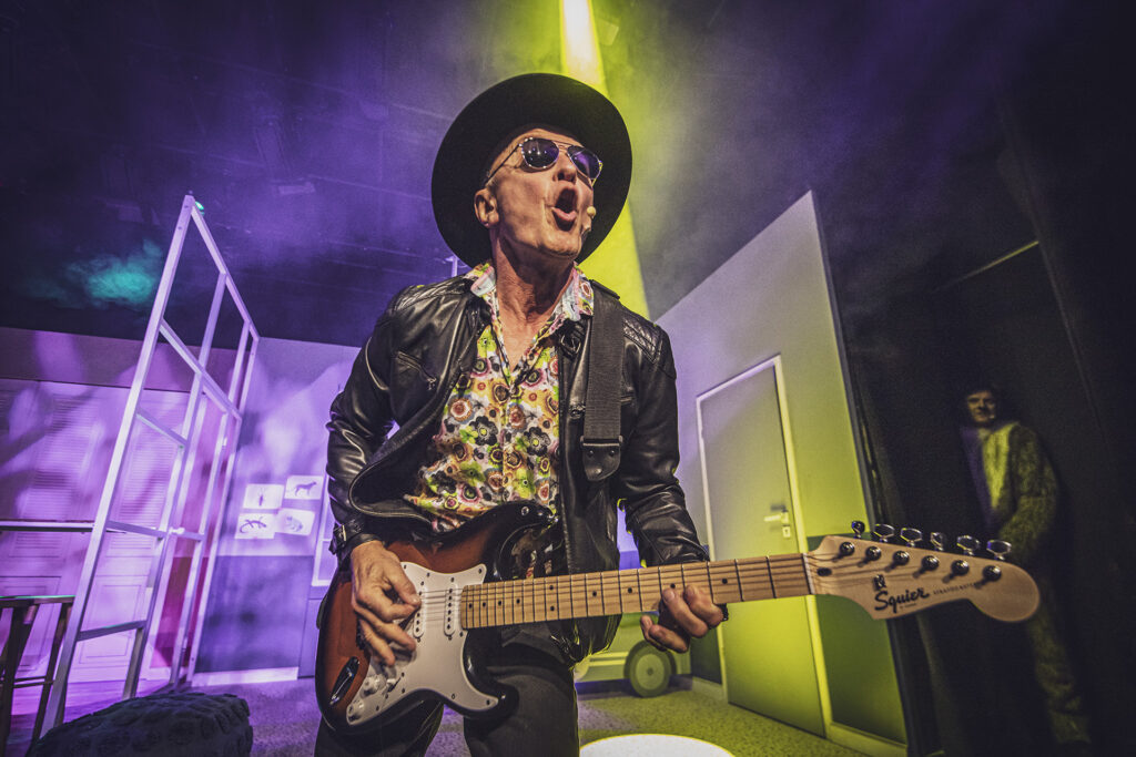 Zdjęcie ze spektaklu Preskot. Aktor w kapeluszu, okularach przeciwsłonecznych i skórzanej kurtce gra na gitarze elektrycznej i śpiewa. Za nim są papierowe ściany mieszkania i drzwi. W tle zza ściany wygląda młody aktor.