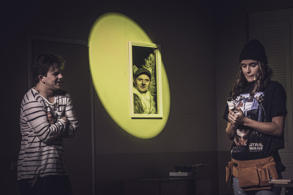 Zdjęcie ze spektaklu Preskot. Młody aktor i aktorka stoją w odstępie. Aktorka ma koszulkę z filmu Star Wars, w ręce trzyma małą figurkę. Między nimi w ramie zawieszonej na ścianie pojawia się aktor w stroju kota.