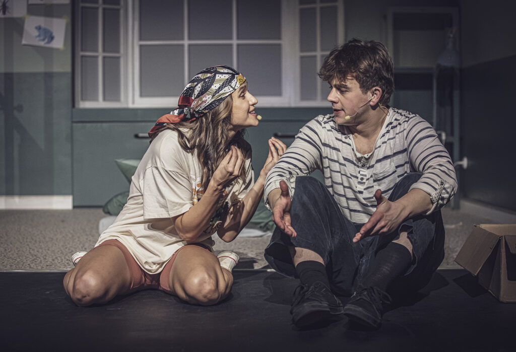 Zdjęcie ze spektaklu Preskot. Młody aktor i aktorka siedzą na podłodze i patrzą na siebie. Aktorka łączy wszystkie palce dłoni i coś intensywnie tłumaczy.