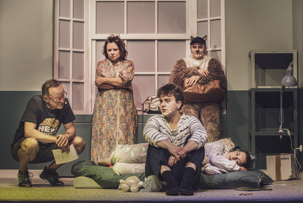 Zdjęcie ze spektaklu Preskot. Młoda aktorka leży na materacu w piżamie. Obok niej siedzi młody aktor. Z boku kuca aktor w średnim wieku w ręce trzyma zapisaną kartkę. Z tyłu aktorka w średnim wieku i aktor w stroju kota opierają się o okno.