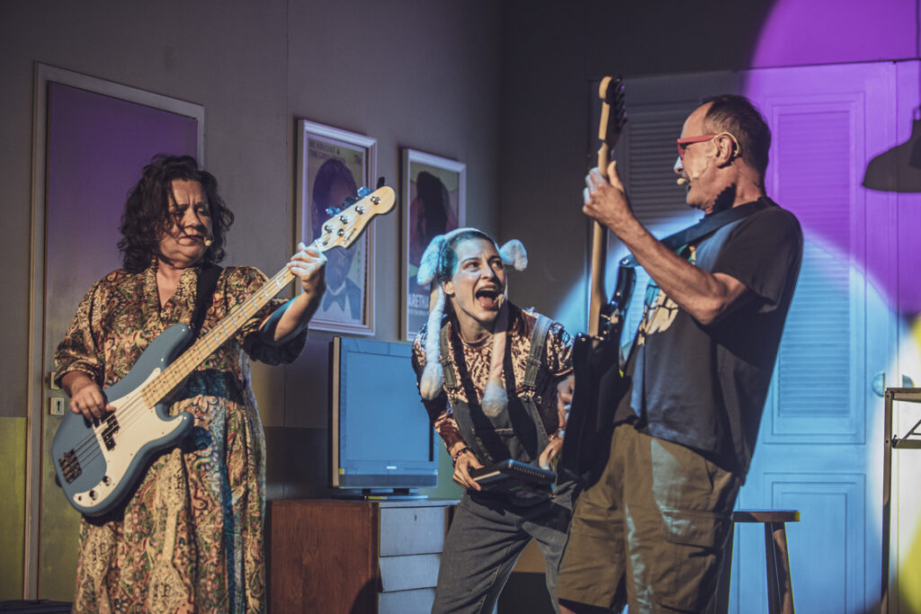 Zdjęcie ze spektaklu Preskot. Aktor i aktorka w średnim wieku grają na gitarach elektrycznych. Między nimi młoda aktorka z uszami królika śpiewa, na szyi ma zawieszony instrument/odtwarzacz muzyczny.