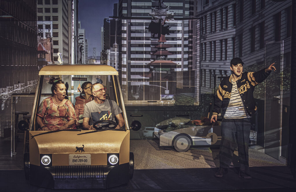 Zdjęcie ze spektaklu Preskot. Aktorka i aktor w średnim wieku oraz młoda aktorka siedzą w makiecie samochodu. Obok stoi młody aktor bejsbolówce, czapce z daszkiem i wskazuje palcem przed siebie. W tle na ścianie wyświetlony jest obraz miasta.