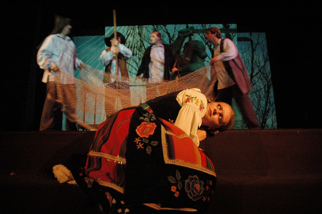 Zdjęcie ze spektaklu Pyza na polskich dróżkach. Na pierwszym planie leży skulona aktorka w łowickim stroju ludowym. W tle czterech aktorów rozciąga sieci rybackie. Na ekranie wyświetlone jest zdjęcie syrenki warszawskiej.