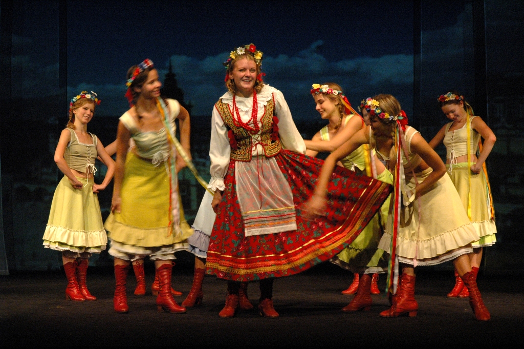 Zdjęcie ze spektaklu Pyza na polskich dróżkach. Scena zbiorowa. Na środku stoi aktorka z warkoczami w krakowskim stroju ludowym. Na głowie ma wianek. Wokół niej tańczą dziewczyny w spódnicach, czerwonych trzewikach i wiankach na głowach.