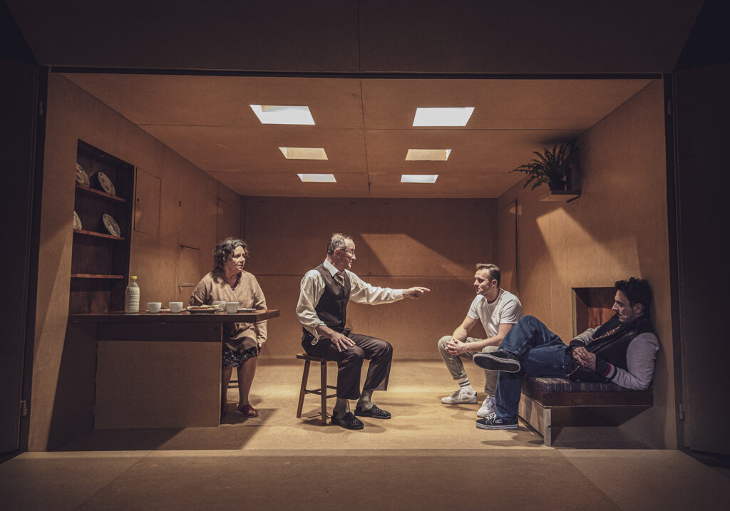 Zdjęcie ze spektaklu Śmierć komiwojażera. Na taborecie siedzi aktor w średnim wieku. Za nim przy stole siedzi aktorka w średnim wieku. Na stole są filiżanki, kanapki i mleko. Z drugiej strony na rozkładanym łóżku siedzi dwóch młodych aktorów.