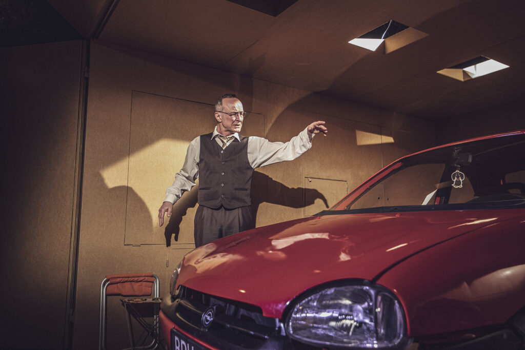 Zdjęcie ze spektaklu Śmierć komiwojażera. Aktor w średnim wieku stoi obok starego opla. Aktor patrzy na samochód i unosi rękę. Aktor ma okulary, koszulę i kamizelkę.