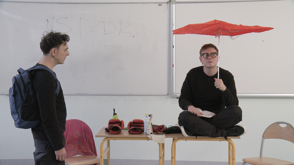 djęcie ze spektaklu Trik Patryka. Aktor w okularach siedzi po turecku na ławce. Na kolanach ma książkę, w ręce trzyma parasol. Na ławce są rękawice i karton mleka. Obok ławki stoi bokiem aktor z plecakiem. W tle jest tablica magnetyczna.