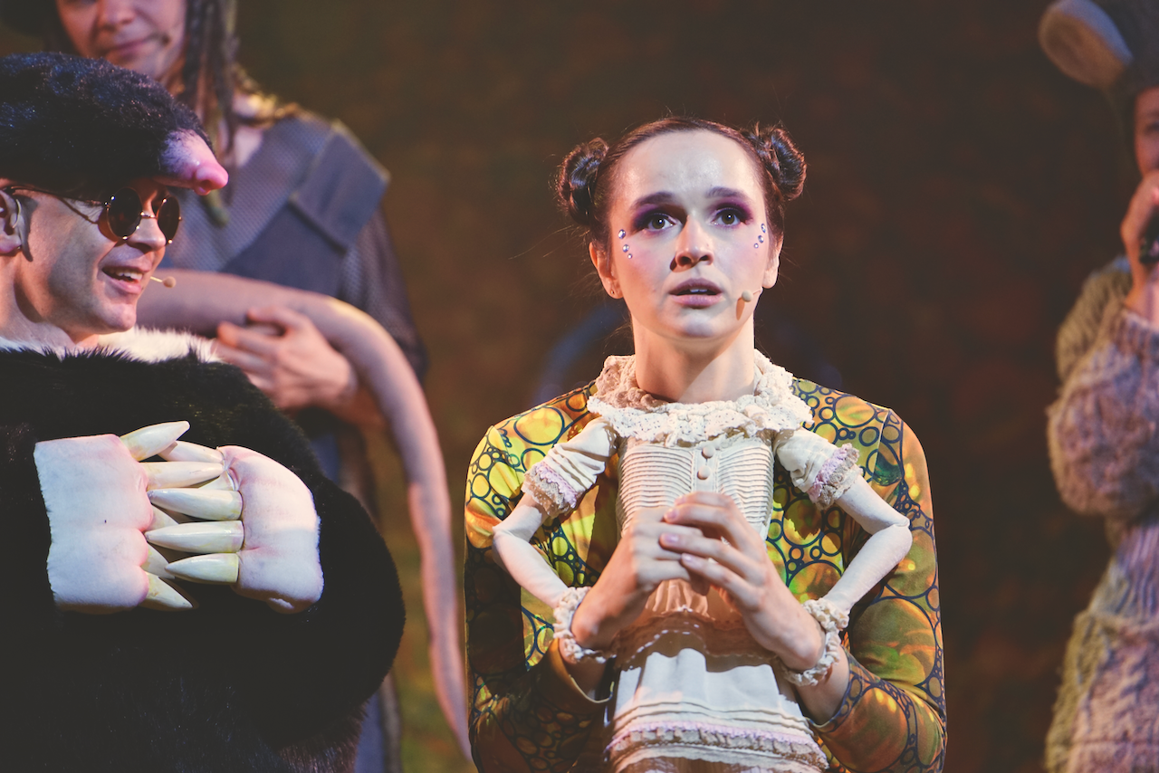 Zdjęcie ze spektaklu Calineczka. Na pierwszym planie aktorka z przyczepiona do kostiumu ciałem małej lalki. Obok niej siedzi aktor przebrany za kreta, czarne futro , czarne okulary.