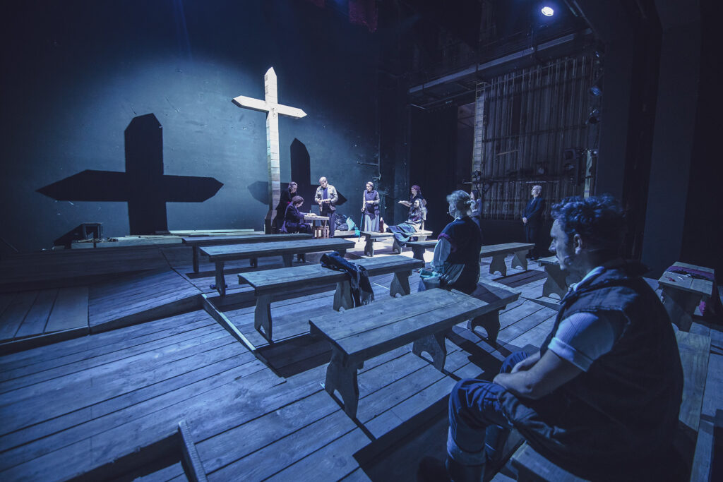 Zdjęcie ze spektaklu Chłopi. Zdjęcie zabarwione na niebiesko. Aktorzy i aktorki w kostiumach wiejskich siedzą na drewnianych ławach i patrzą w stronę drewnianego krzyża. Kilka osób stoi pod krzyżem.