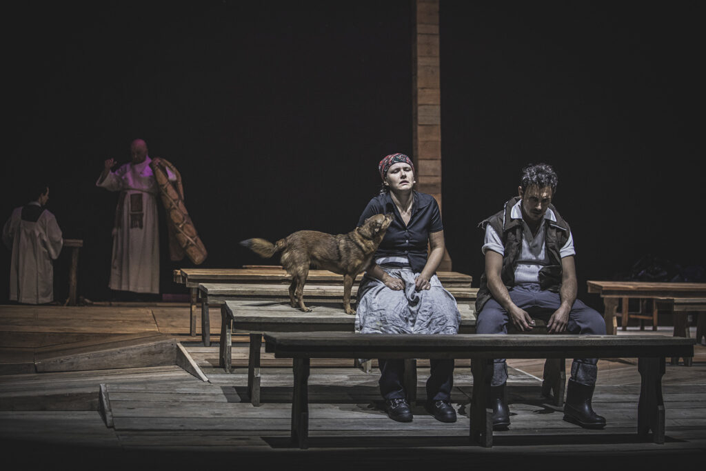 djęcie ze spektaklu Chłopi. Na drewnianej ławie siedzi aktorka i aktor w wiejskich strojach. Obok kobiety na ławce stoi pies. W tle stoi aktor w stroju księdza i aktor w stroju kościelnego.