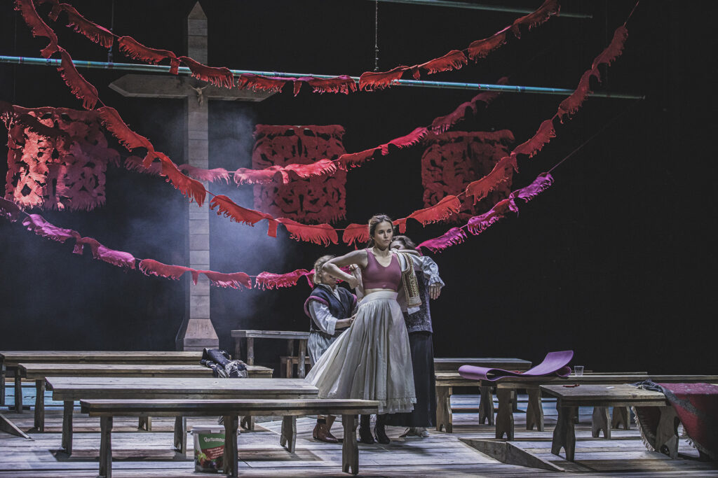 Zdjęcie ze spektaklu Chłopi. Na środku stoi aktorka. Dwie aktorki stoją za nią i ubierają ją w strój ludowy. Na scenie są drewniane ławy i drewniany krzyż jak w wiejskim kościele. Z sufitu zwisają wycinanki ludowe