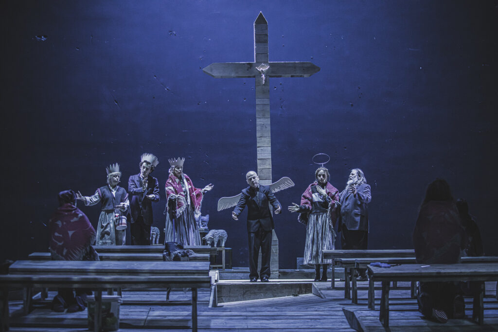 Zdjęcie ze spektaklu Chłopi. Scena zbiorowa. Pod krzyżem stoją aktorzy i aktorki w wiejskich strojach. Na głowach mają korony. Jedna aktorka na głowie ma aureolę. Jeden aktor ma skrzydła na plecach. Za nimi stoją figurki baranków.