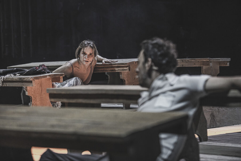 Zdjęcie ze spektaklu Chłopi. Aktorka i aktor kucają między drewnianymi ławami. Patrzą na siebie.