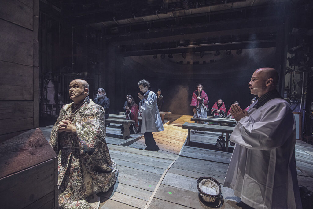 Zdjęcie ze spektaklu Chłopi. Scena zbiorowa. Aktor w stroju księdza klęczy pod krzyżem. Obok dwóch aktorów w strojach kościelnych. Na podłodze leży koszyk na ofiarę. W tle na drewnianych ławach siedzą aktorzy i aktorki w ludowych chustach.