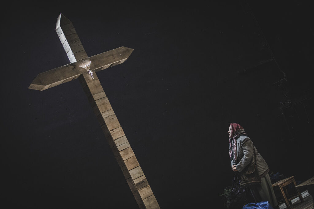Zdjęcie ze spektaklu Chłopi. Pod drewnianym krzyżem stoi starsza kobieta w chuście.
