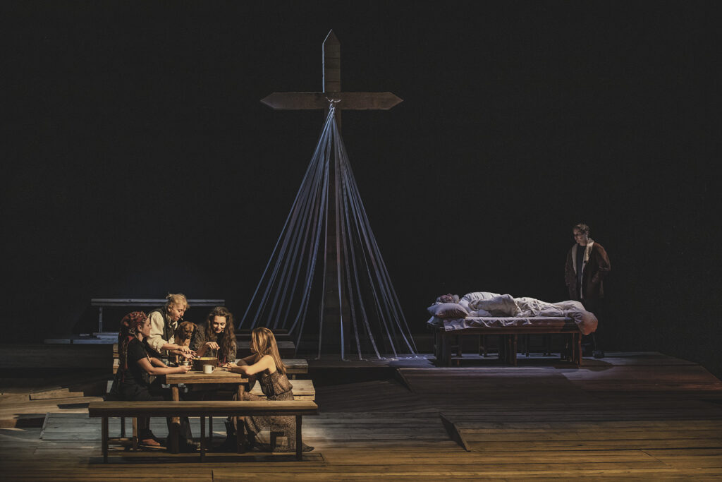 Zdjęcie ze spektaklu Chłopi. Przy stole siedzą cztery aktorki w wiejskich strojach, między nimi jest pies. Z tyłu na łóżku leży starszy aktor, obok niego stoi młody aktor. Nad nimi jest niebieskie światło. W tle jest drewniany krzyż z wstążkami.