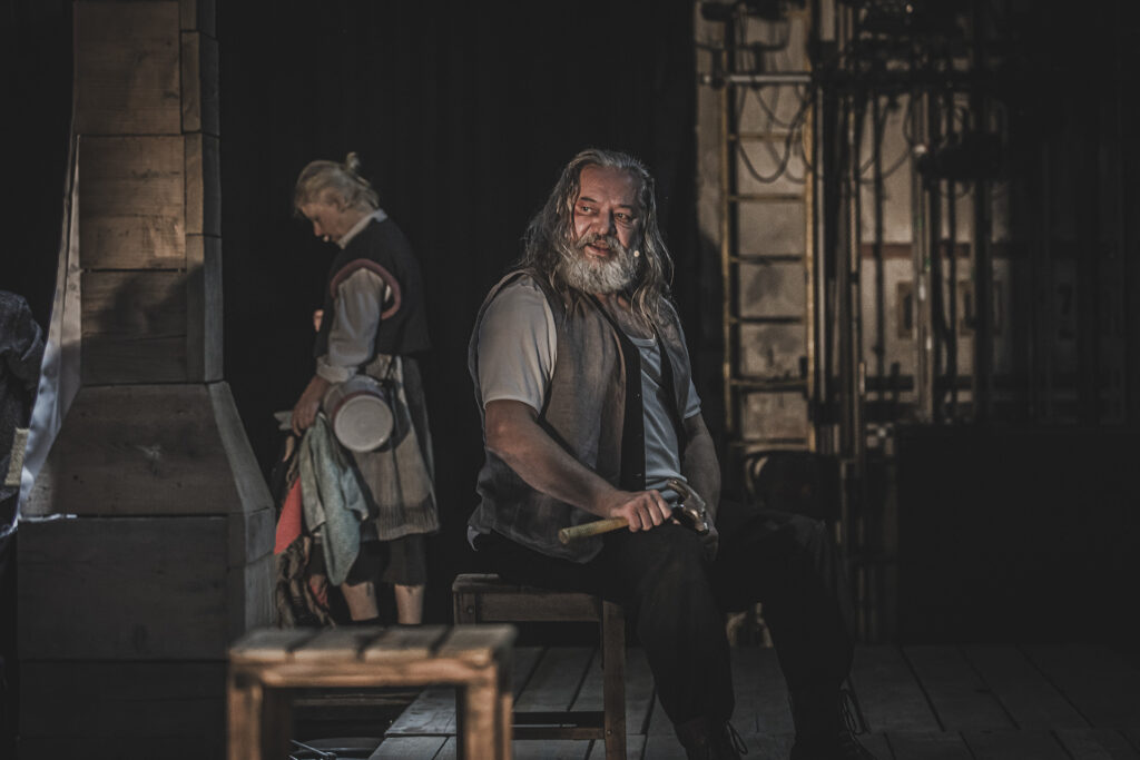 Zdjęcie ze spektaklu Chłopi. Starszy aktor z długimi włosami i brodą siedzi na drewnianym stołku. W ręce trzyma młotek. W tle stoi aktorka w wiejskim stroju.