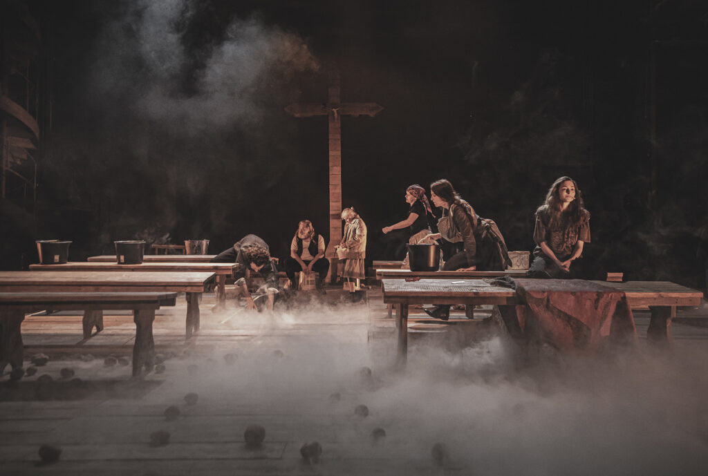 Zdjęcie ze spektaklu Chłopi. Scena zbiorowa. Aktorzy i aktorki w wiejskich strojach siedzą na drewnianych ławach lub stołkach. Na środku sceny jest drewniany krzyż. Na ławach stoją wiadra. Na podłodze są rozsypane ziemniaki. Scenę spowija mgła.