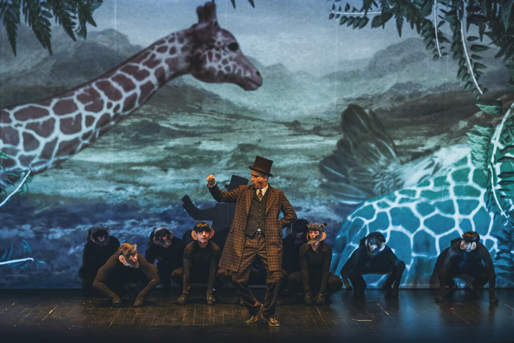 Zdjęcie ze spektaklu Doktor Dolittle. Aktor w staromodnym garniturze i cylindrze tańczy na środku. Wokół niego kuca grupa aktorów w kostiumach małp. Mają czarne peruki, okulary i doklejone uszy. Na ścianie w tle jest wyświetlony obraz żyrafy.
