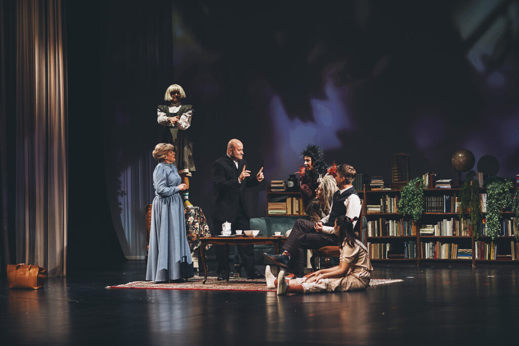 Zdjęcie ze spektaklu Doktor Dolittle. Scena zbiorowa. Aktorzy i aktorki siedzą lub stoją przy ławie. Niektórzy mają peruki oznaczające kostiumy zwierząt. Za nimi w tle jest mała biblioteczka.