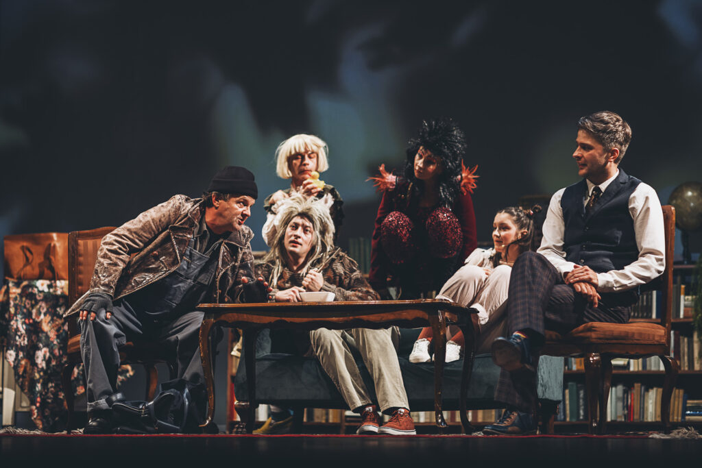 Zdjęcie ze spektaklu Doktor Dolittle. Na kanapie siedzi dwóch aktorów rozmawiają. Wokół nich siedzą aktorzy w strojach wróbla, papugi, kaczki i świnki.