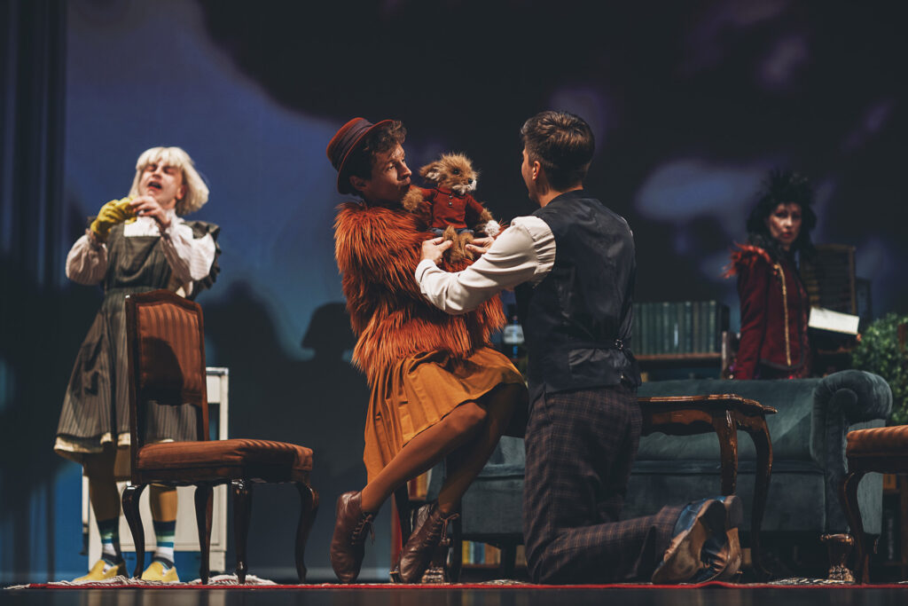 Zdjęcie ze spektaklu Doktor Dolittle. Aktor w rudym futrze i kapeluszu siedzi na ławie i trzyma lalkę lisa. Przed nim klęczy aktor w garniturze. Z boku stoi aktor w stroju kaczki. W tle stoi aktorka w stroju papugi.