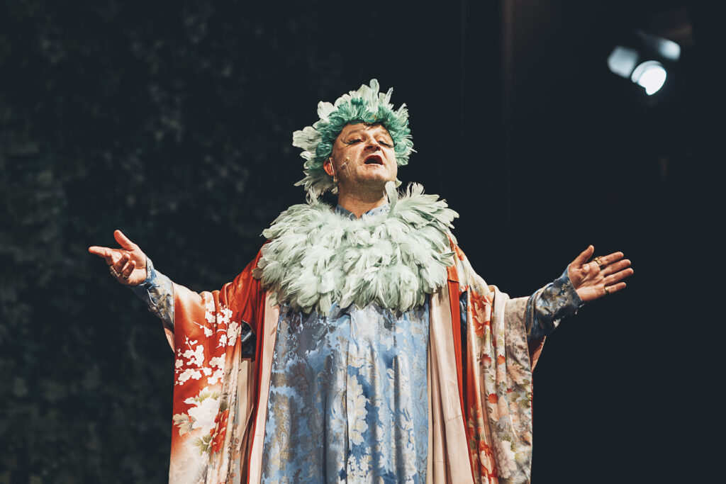 Zdjęcie ze spektaklu Doktor Dolittle. Aktor w stroju kanarzycy. Ma kolorowy strój z piórami. Unosi ręce i śpiewa.