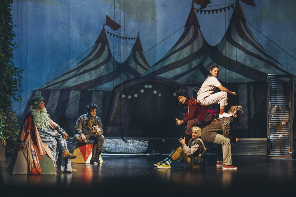 Zdjęcie ze spektaklu Doktor Dolittle. Scena zbiorowa. Aktorzy w strojach kanarzycy i wróbla patrzą na piramidę z ciał aktorów i aktorek w strojach świnki, papugi, psa i kaczki. Z boku w klatce siedzi aktor. Na ścianie wyświetlony jest obraz cyrku.