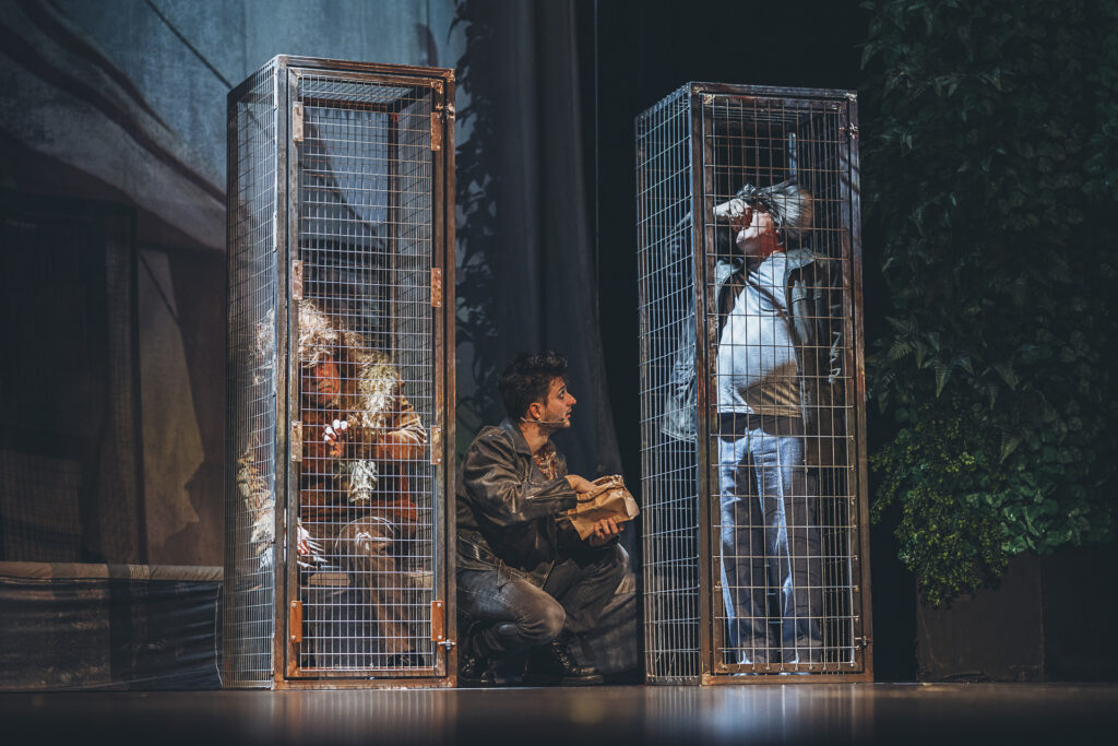 Zdjęcie ze spektaklu Doktor Dolittle. W dwóch dużych pionowych klatkach siedzą aktorzy w stroju zwierząt. Między klatkami kuca młody aktor, w ręce ma papierową torbę.