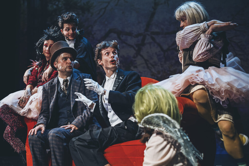 Zdjęcie ze spektaklu Doktor Dolittle. Na kanapie siedzi dwóch aktorów w staromodnych garniturach. Wokół nich siedzą aktorzy w strojach wróbla, papugi, kaczki i świnki.