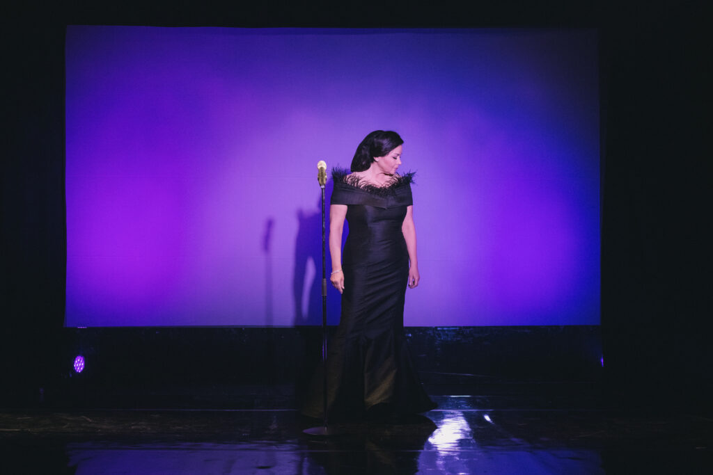 Zdjęcie ze spektaklu Głos ludzki/Piaf. Aktorka w długiej czarnej sukni z piórami stoi na scenie. Przekrzywia głowę w bok. Obok niej stoi mikrofon na statywie. Za nią jest ekran z fioletowym tłem. Na ekranie odbija się cień kobiety.