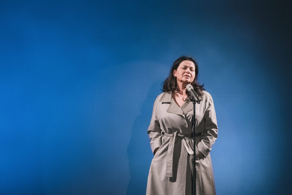 Zdjęcie ze spektaklu Głos ludzki/Piaf. Aktorka w płaszczu stoi przed mikrofonem. Zamyka oczy i trzyma ręce w kieszeniach. Za nią jest niebieskie tło.