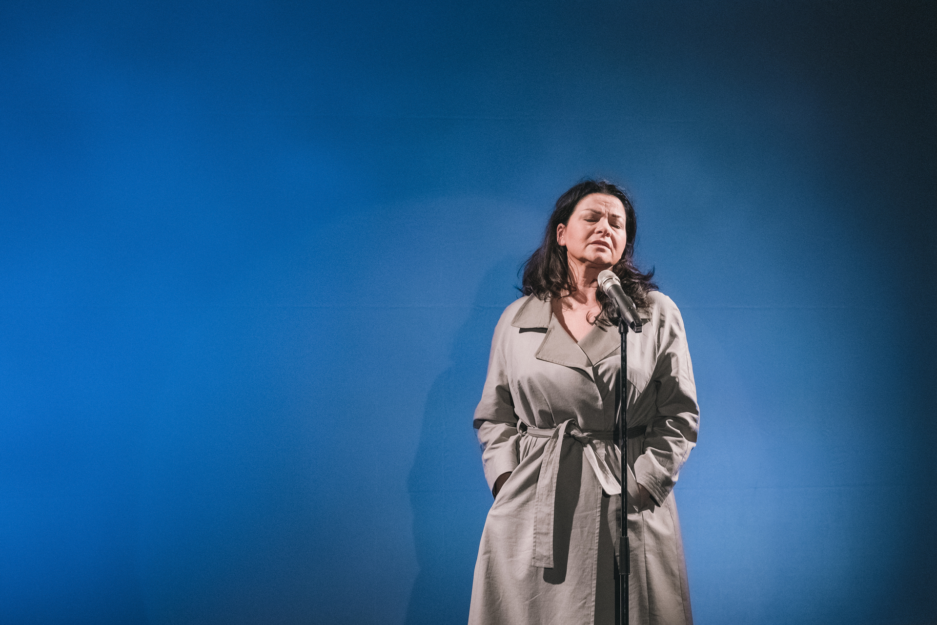 Zdjęcie ze spektaklu Głos ludzki/Piaf. Aktorka w płaszczu stoi przed mikrofonem. Zamyka oczy i trzyma ręce w kieszeniach. Za nią jest niebieskie tło.