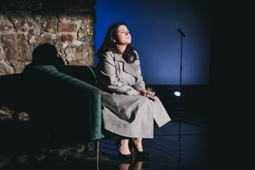 Zdjęcie ze spektaklu Głos ludzki/Piaf. Aktorka siedzi na fotelu. Ma na sobie długi płaszcz i szpilki. W tle jest kamienna ściana, niebieski ekran i mikrofon.