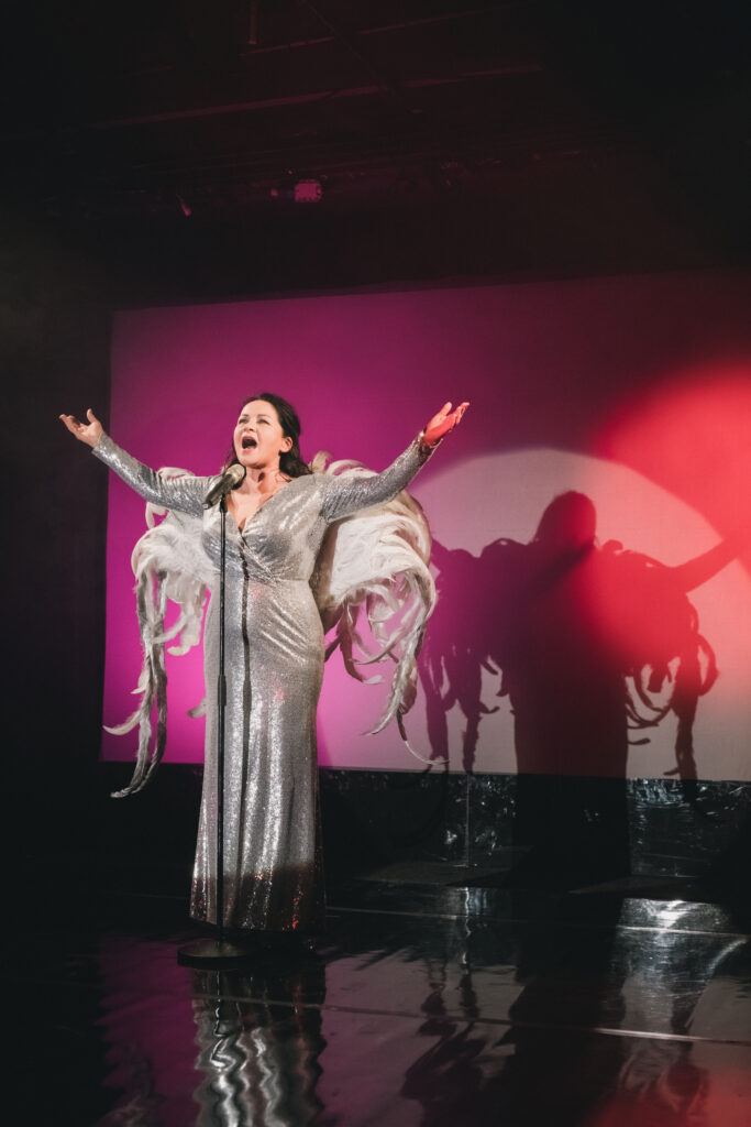 Zdjęcie ze spektaklu Głos ludzki/Piaf. Aktorka stoi przed mikrofonem na statywie, szeroko otwiera usta i unosi ręce. Aktorka ma na sobie długą błyszczącą suknię i skrzydła na plecach. Na czerwonym ekranie w tle odbija się jej cień