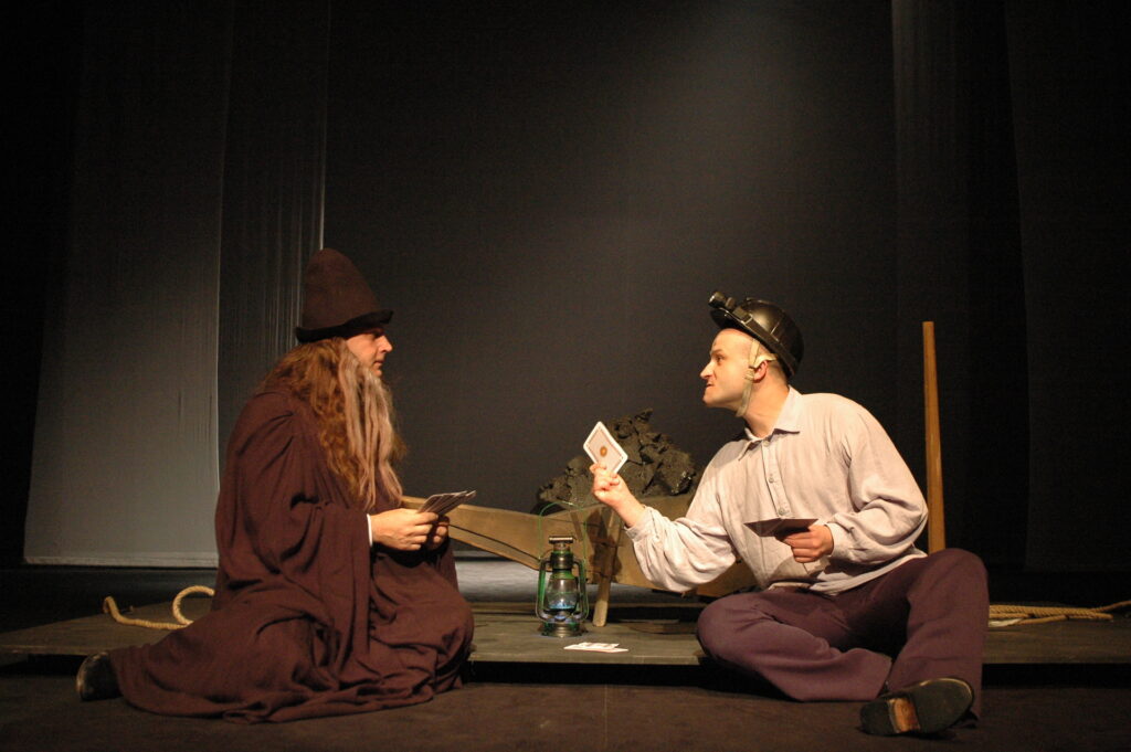 Zdjęcie ze spektaklu Pyza na polskich dróżkach. Aktor w stroju górnika i aktor w stroju skarbnika kopalni siedzą na podłodze i grają w karty. Za nimi stoi lampa górnicza i taczki z węglem.
