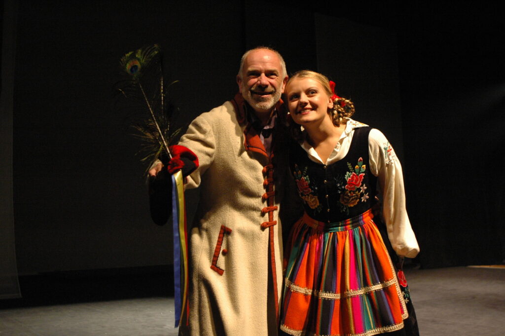 Zdjęcie ze spektaklu Pyza na polskich dróżkach. Młoda aktorka i starszy aktor stoją blisko siebie uśmiechnięci. Mają łowickie stroje.