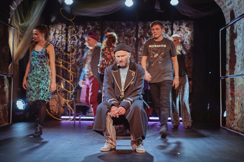 Zdjęcie ze spektaklu Krakowska lekkość bytu. Scena zbiorowa. Na środku na krześle siedzi mężczyzna w długim płaszczu i czapce. Za nim pozostali aktorzy i aktorki chodzą w różnych kierunkach.