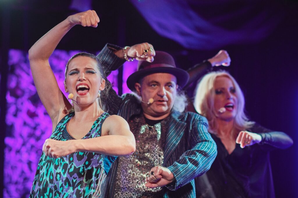 Zdjęcie ze spektaklu Krakowska lekkość bytu. Zbliżenie. Dwie aktorki i aktor tańczą i śpiewają. Aktor jest w środku. Wszyscy mają dłonie zaciśnięte w pięści, jedną rękę trzymają na wysokości klatki piersiowej, a drugą nad głową.