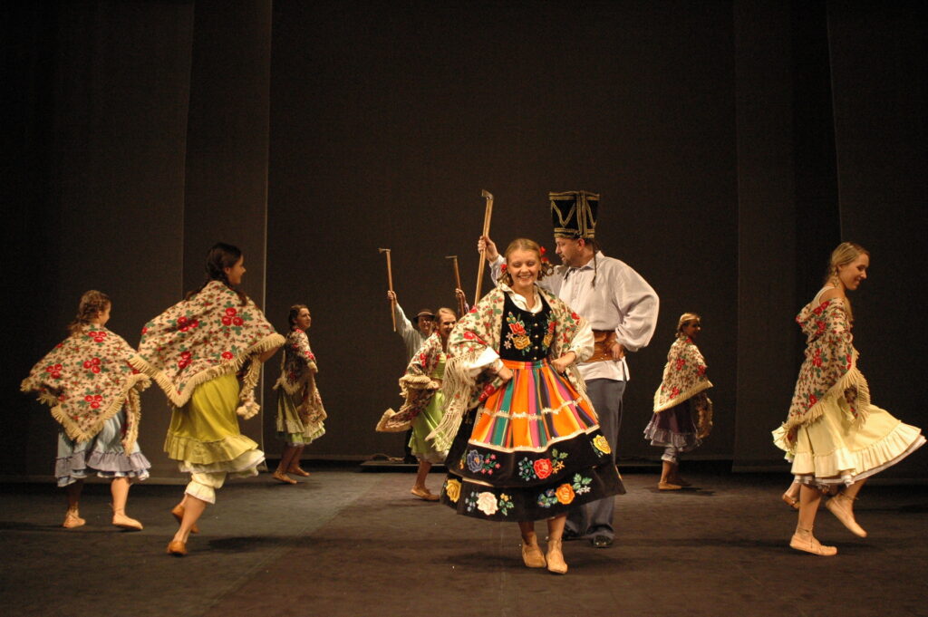 Zdjęcie ze spektaklu Pyza na polskich dróżkach. Scena zbiorowa. Kobiety w spódnicach, kierpcach i góralskich chustach na ramionach tańczą w koło. Między nimi tańczą mężczyźni w czapkach i z ciupagami.