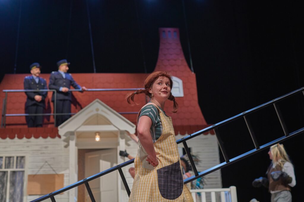 Zdjęcie ze spektaklu Pippi. Aktorka trzyma drabinę. Ma bluzkę w paski, sukienkę w kratkę i dwa warkocze sterczące na boki. W tle jest jej domek. Obok jest aktor w stroju konia. Na dachu są aktorzy w strojach policjantów.