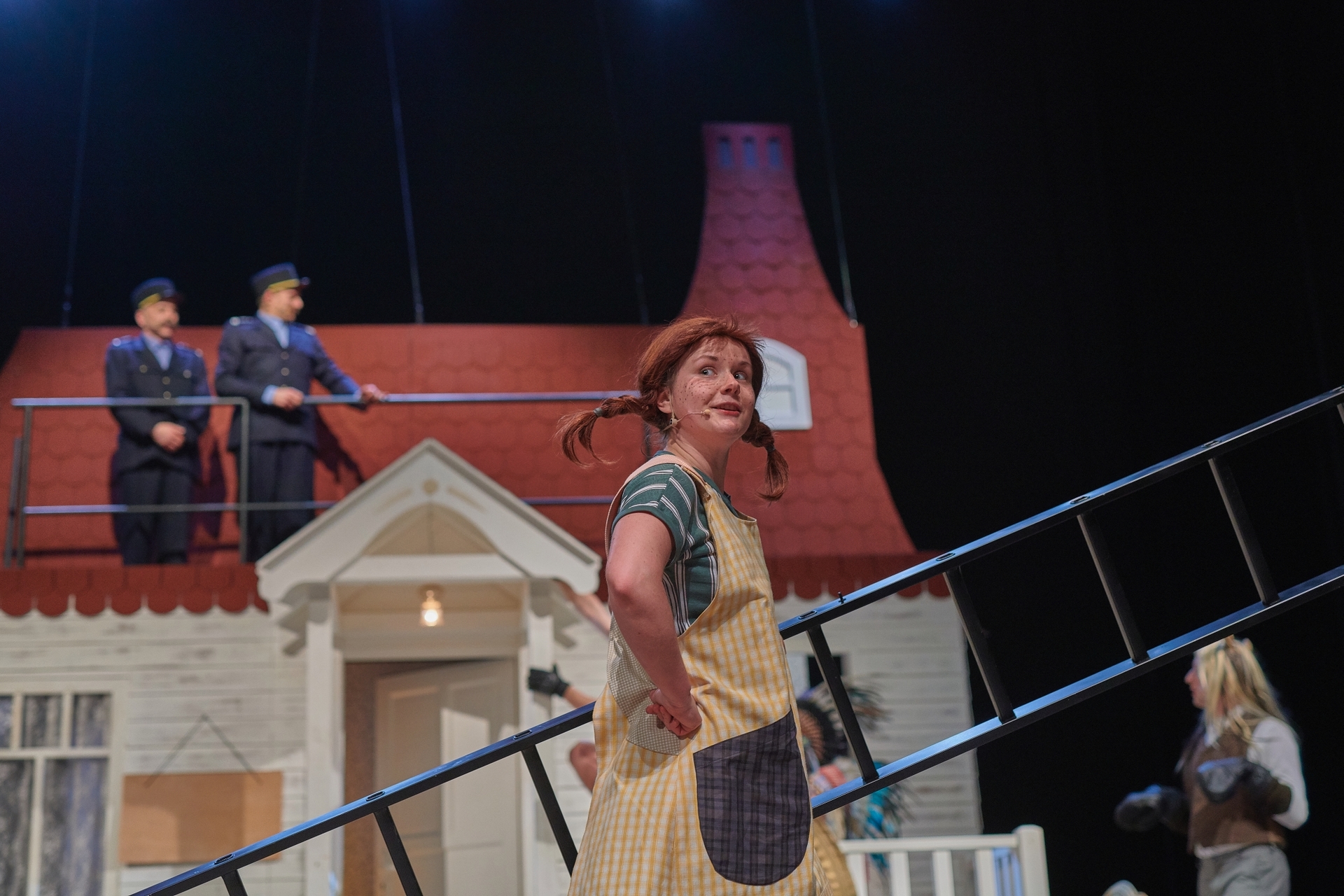 Zdjęcie ze spektaklu Pippi. Aktorka trzyma drabinę. Ma bluzkę w paski, sukienkę w kratkę i dwa warkocze sterczące na boki. W tle jest jej domek. Obok jest aktor w stroju konia. Na dachu są aktorzy w strojach policjantów.