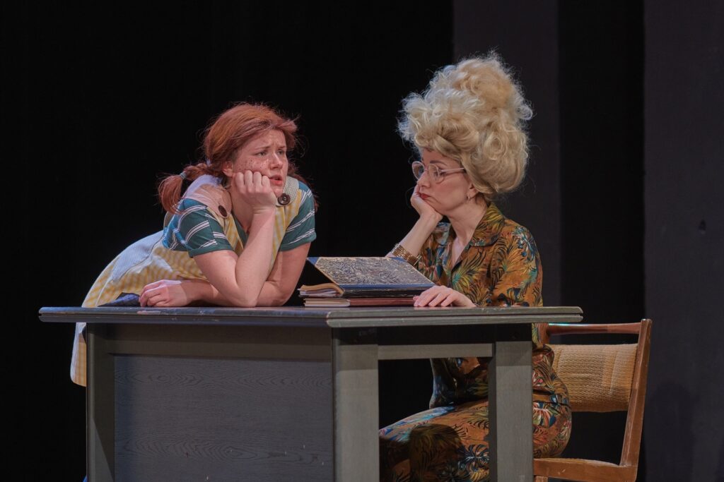 Zdjęcie ze spektaklu Pippi. Przy biurku siedzi aktorka w okularach i natapirowanej fryzurze. Podpiera brodę dłonią. Obok na biurku opiera się aktorka z warkoczami w stroju Pippi. Też podpiera brodę dłonią. Na stole leżą zeszyty.