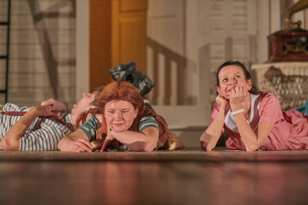 Zdjęcie ze spektaklu Pippi. Aktorka z warkoczami leży na dywanie, rękę trzyma pod brodą, ma splecione nogi. Aktor w koszulce w paski opiera się o jej plecy. Obok leży aktorka w sukience w kratkę.