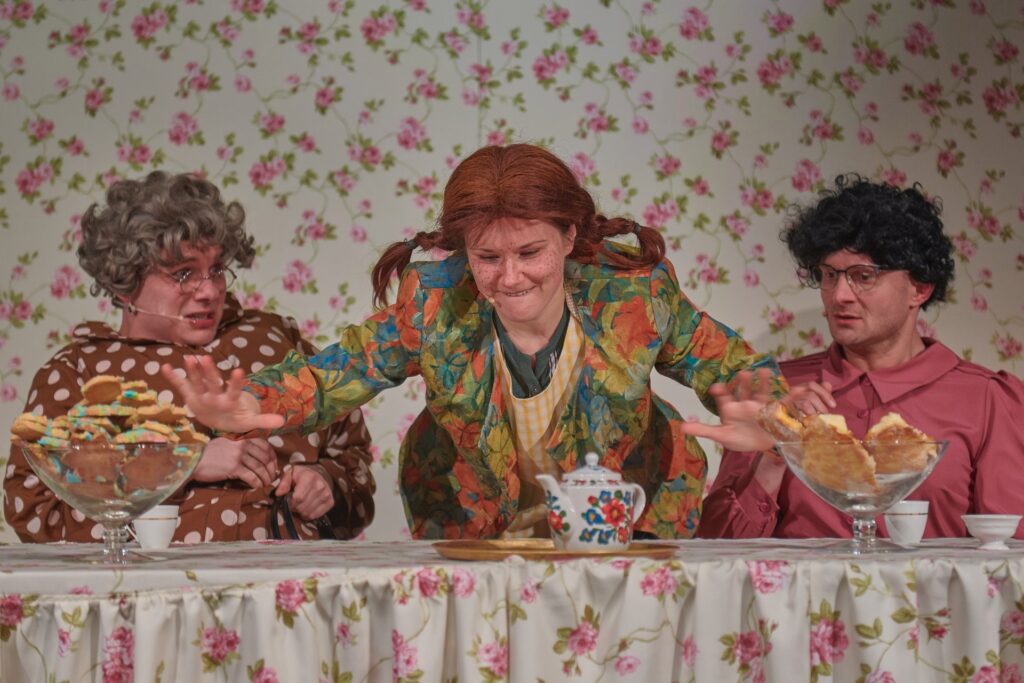 Zdjęcie ze spektaklu Pippi. Aktorka z warkoczami pochyla się nad stołem z obrusem w kwiaty i dotyka ciastek. Obok siedzą aktorzy przebrani za kobiety. Mają sukienki, peruki i okulary. W tle jest tapeta w kwiaty takie jak na obrusie.