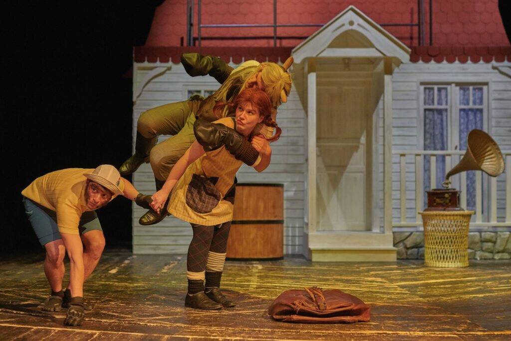 Zdjęcie ze spektaklu Pippi. Aktorka z warkoczami niesie na plecach aktora w stroju konia. Obok nich jest pochylony aktor w stroju małpy. W tle jest mały domek. Przed domem na stoliku stoi gramofon.