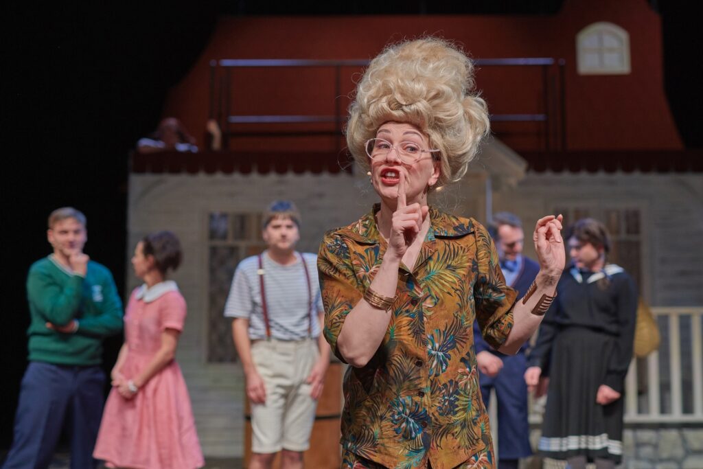 Zdjęcie ze spektaklu Pippi. Aktorka w okularach i natapirowanej fryzurze otwiera usta i unosi palec wskazujący. Za nią w rzędzie stoją aktorzy i aktorki w strojach uczniów. W tle jest mały domek.