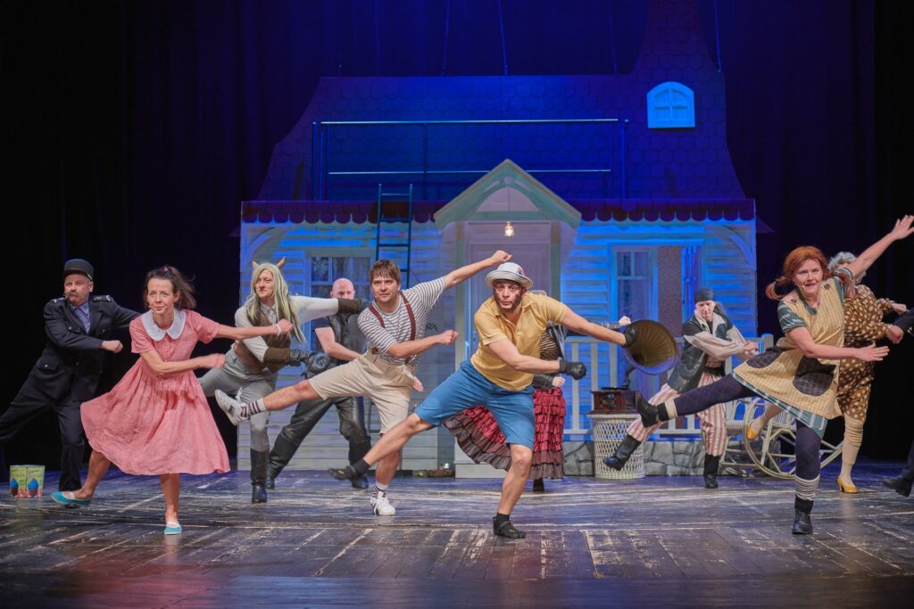 Zdjęcie ze spektaklu Pippi. Scena zbiorowa. Wszyscy aktorzy i aktorki tańczą. Stoją na jednej nodze, drugą unoszą do góry i machają rękami. W tle jest mały domek.