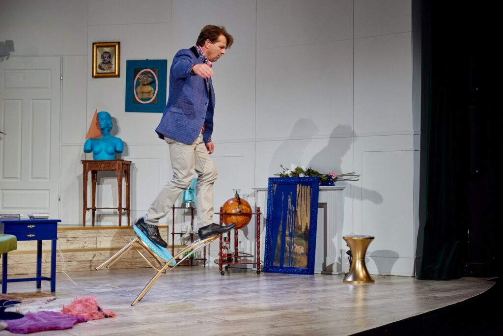 Zdjęcie ze spektaklu Kolacja dla głupca. Aktor w eleganckim stroju stoi na krześle, które się przewraca. Jedną nogę trzyma na siedzeniu, a drugą na oparciu krzesła. W tle jest mieszkanie z awangardowymi dziełami sztuki.